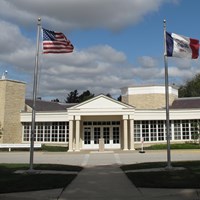 A sprawling one story public building of rough-faced yellowish stone has a white portico entrance.