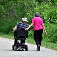A woman walks beside a man on a flat trail, resting a hand on the back of his motorized wheelchair.