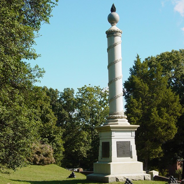 A rounded white cement type pillar stands above a green field. A brass plaque at the base. 