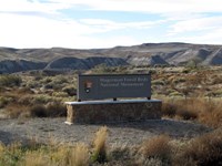 A brown sign introducing the park entrance with large eroded brown hills behind it.