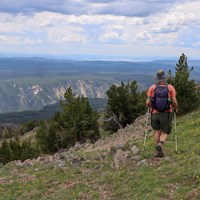 A hiker walks on a mountain slope with a canyon and lake seen in the distance.