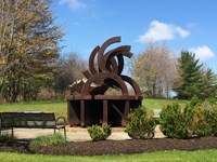 Rust-colored metal sculpture depicting broken Olympic rings surrounded by a circular cement walkway.