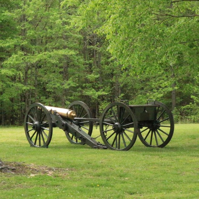 A cannon and Limber stand on a green lawn between trees. A stand of trees is on the horizon. 