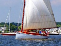 A sailboat with a large white sail on a calm body of water near a marina, under a partly cloudy sky.