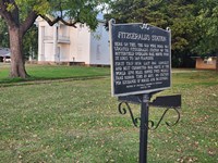A blue sign sits on the lawn in front of a large white colonial house.