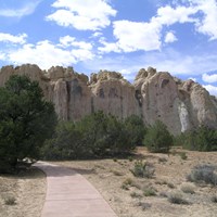 A trail winds through juniper trees towards the base of a large cliff.