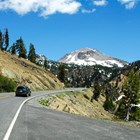 A black car on a winding mountain road lined by steep slopes with a large volcano.