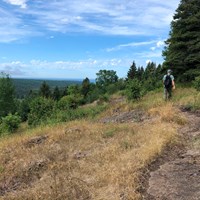 A person hikes along a trail on a ridge top. Trees cover the rolling landscape.