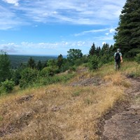 A person hikes along a trail on a ridge top. Trees cover the rolling landscape. 