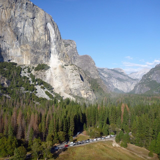 A large granite mountain towers over a parking lot full of cars