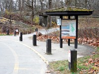 Three sided kiosk beside parking lot posts. A paved path leads to a stone lock and wooded trail.