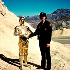A park ranger stands next to a gold robot, shaking hands, in the desert. 