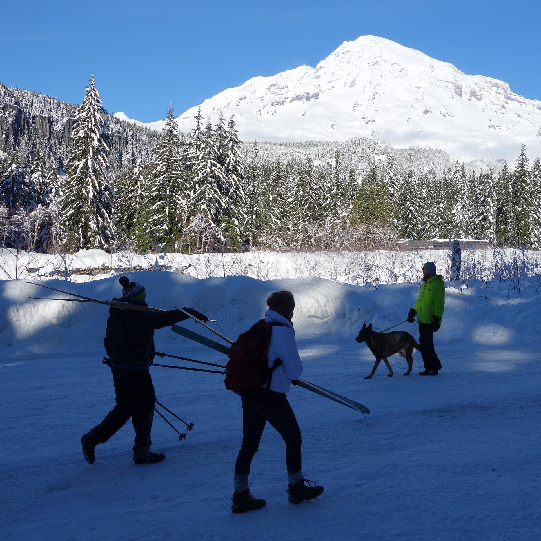 Several people walk along a snowy road in front of a view of a snow-covered mountain and forest.