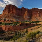 Prominent red cliffs with blue sky, clouds in distance, with red slopes and green grass in front.