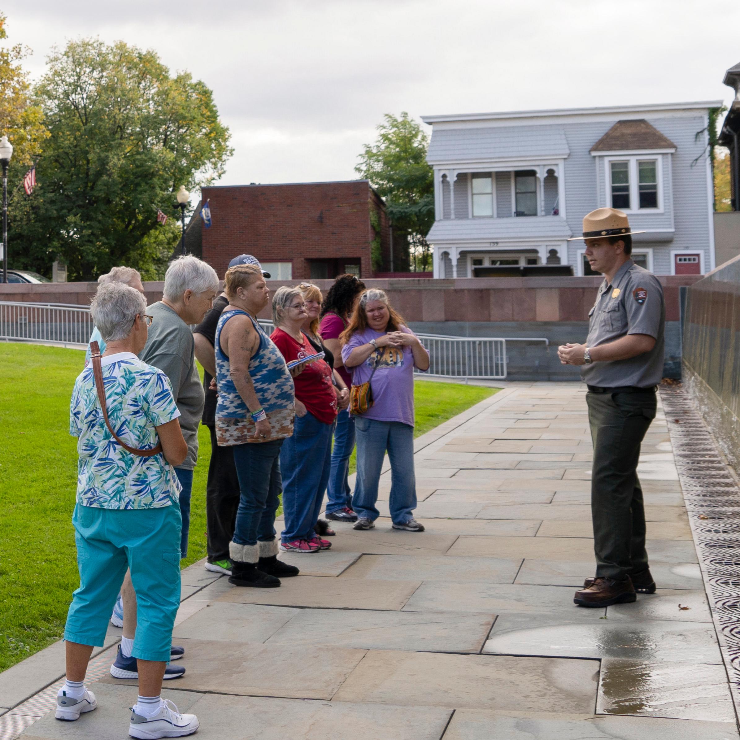 A ranger stands and speaks to a group of people in front of a wall.