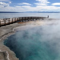 A turquoise hot spring pool surrounded by a boardwalk sits above a large lake.