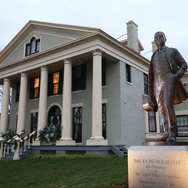 A life-sized bronze statue of TR with a large white columned house behind it.