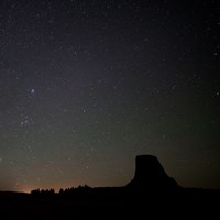 a dark night sky filled with stars with Devils Tower and trees silhouetted below