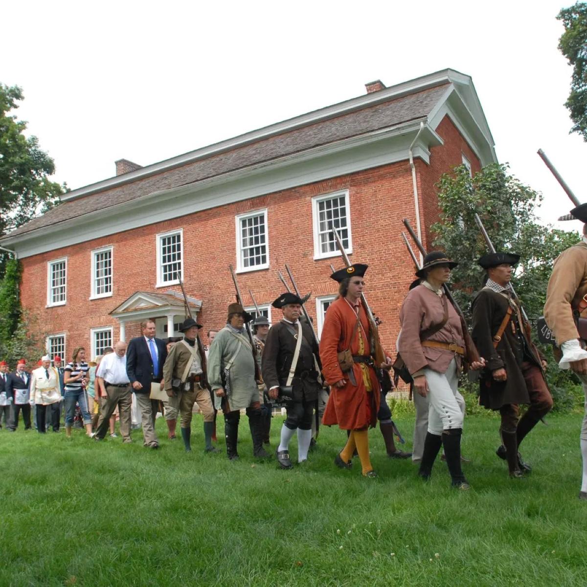 A line of people dressed as 18th C militia and in modern clothes march past a large brick house.