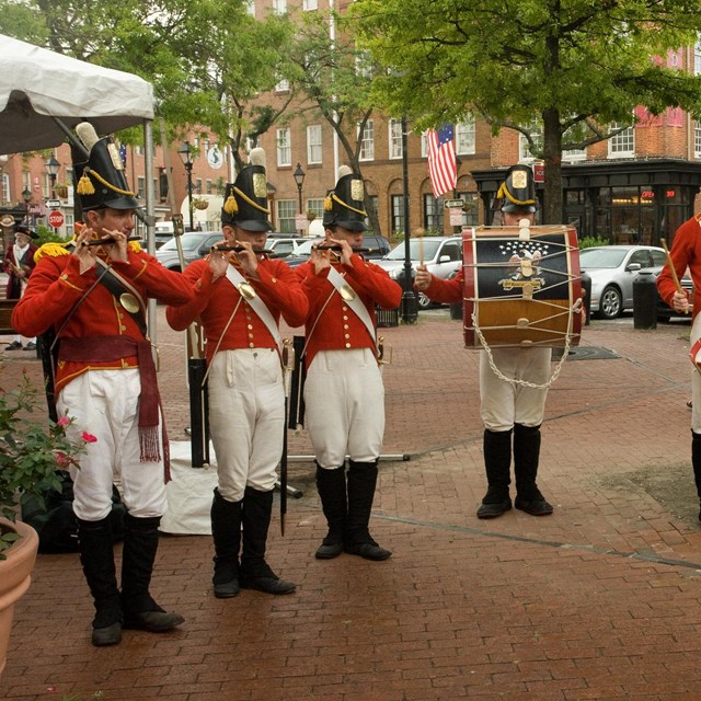 Several soldiers with fifes and drums stand in a line with red jackets. They have large black hats. 