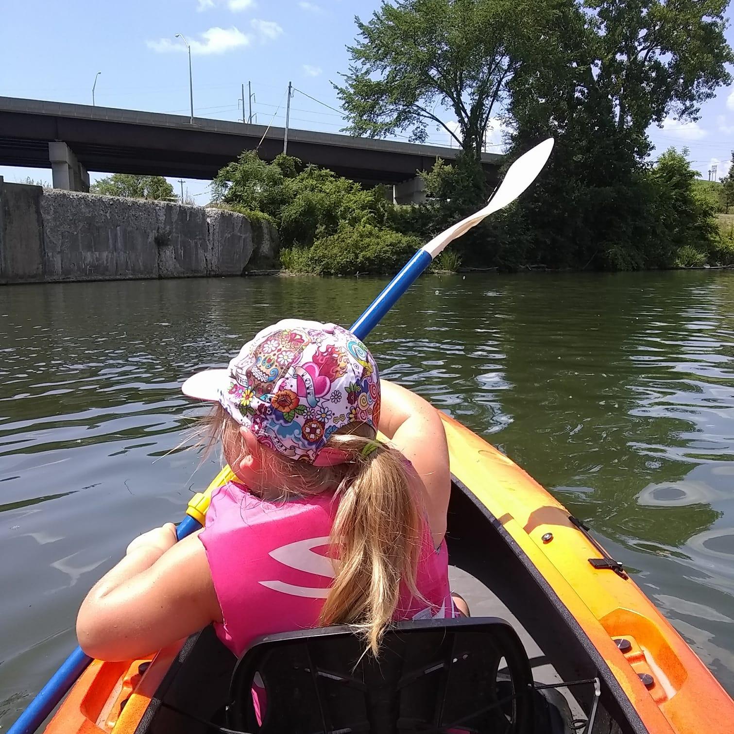 You are seated behind a little girl with a ponytail. You are in a kayak and she is holding a paddle.