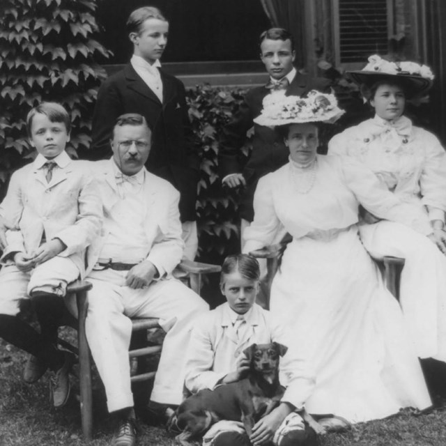 Black & white a family in their finest sits. 4 sons, 1 daughter, mother, and father Teddy Roosevelt.