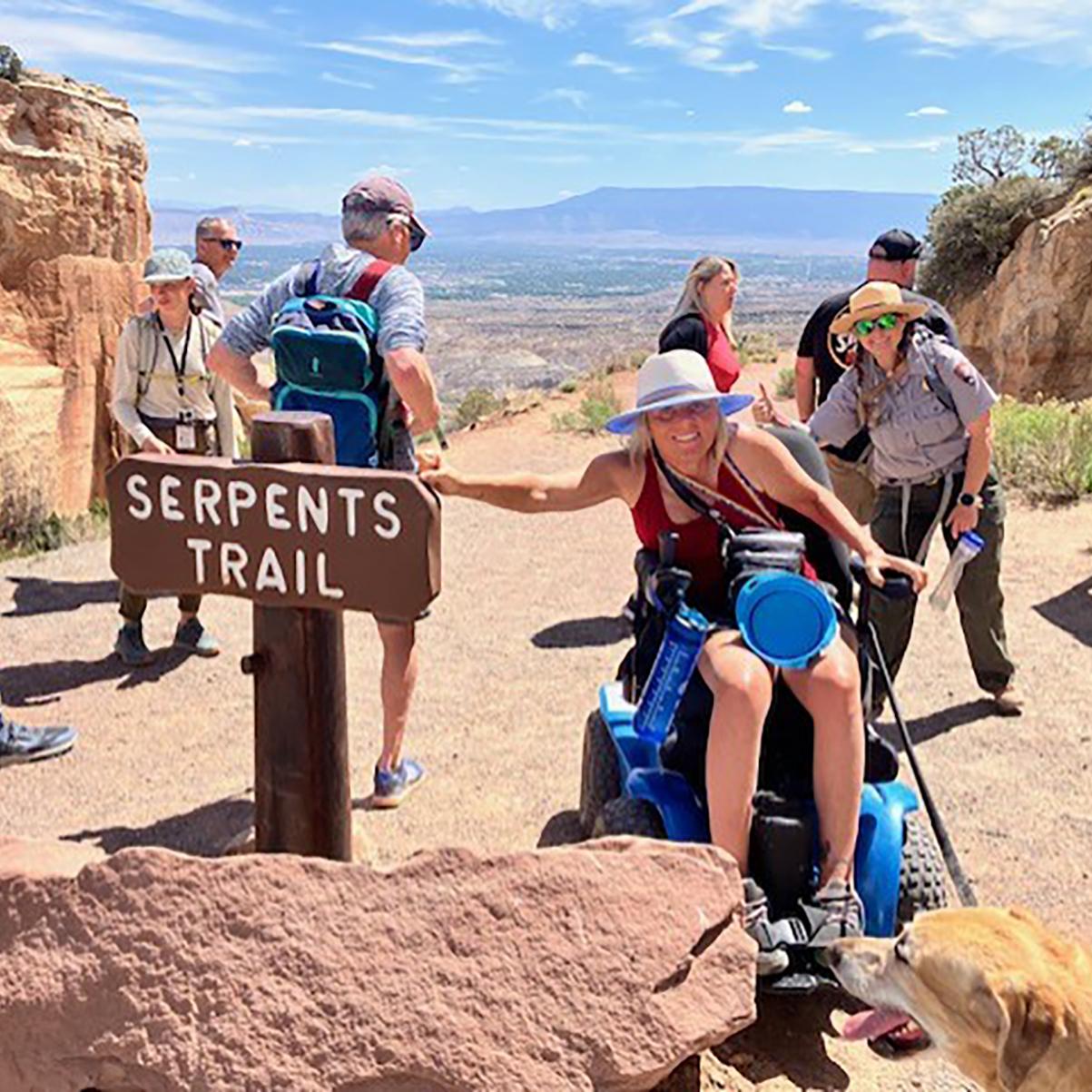 woman using wheelchair and service animal points at Serpents Trail sign with uniformed ranger behind