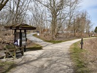 A three-sided kiosk and bench are at a fork. The trail on the right is beside a water-filled canal.