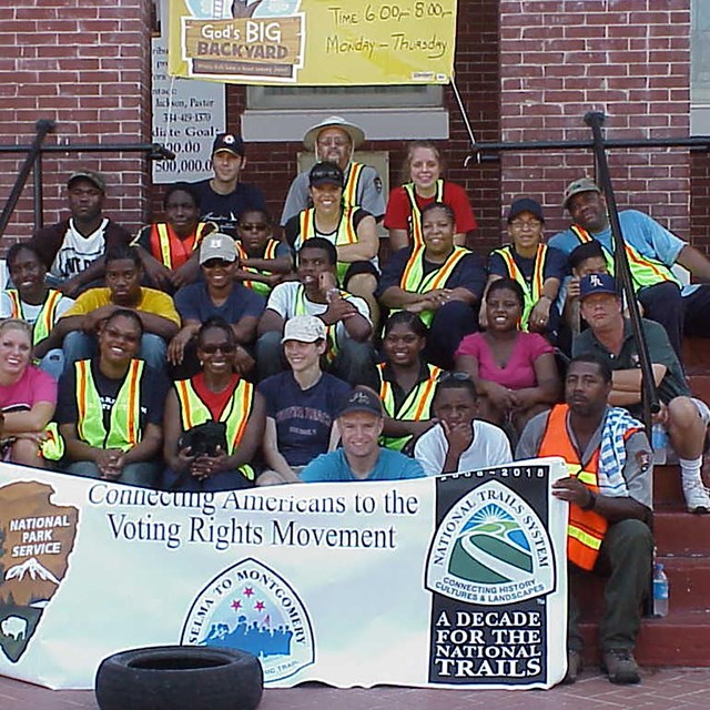 Large group of diverse people sitting on stone steps holding a trails clean up banner.