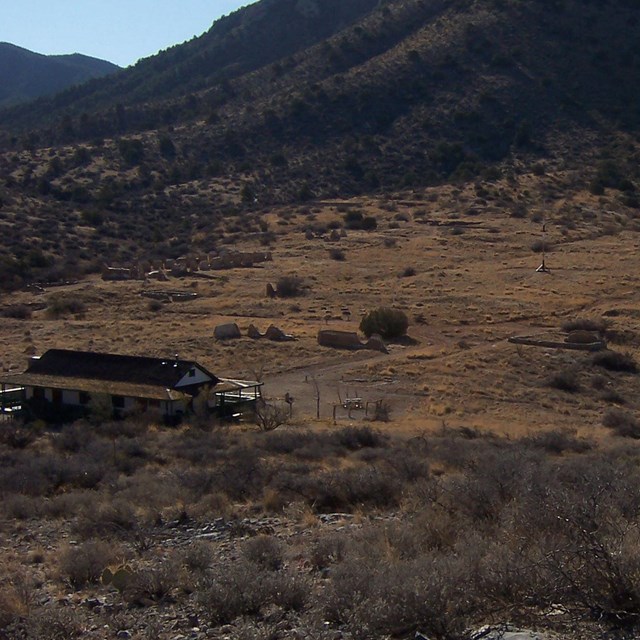 A large 1-story building sits in a grassy field surrounded by tall, rocky mountain sides.