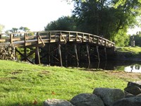 Arched wooden bridge with field stone abutments spans a slow river bounded by green embankments