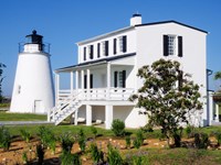 Piney Point Lighthouse and adjacent white brick building with gardens, under a clear blue sky.
