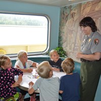 Children sit around a table working on Junior Ranger booklets while a Park Ranger stands nearby.