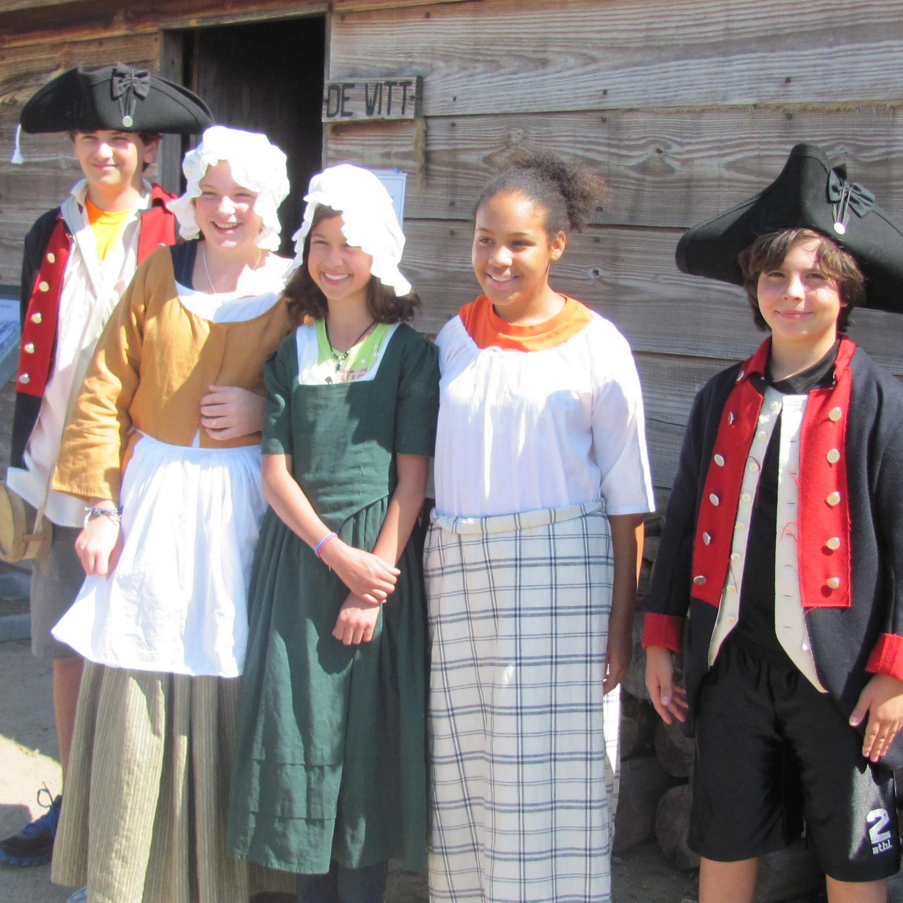 Five children stand in a row smiling. They all have different 18th Century accessories on.