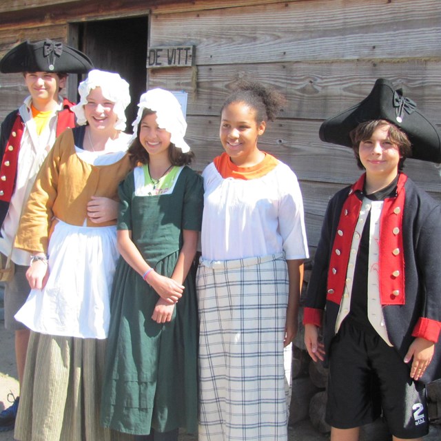 Several children wear colonial style clothing in many colors. Wooden buildings sit behind them.