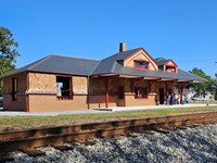 An historic brick and wood train station with slate-gray roof and a group of people on the platform.