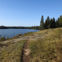 A trail by a small hill that leads to a lake. Forest in the distance.