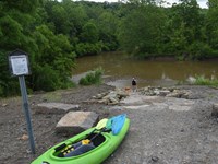 A green kayak rests by a trail sign overlooking the junction of two waterways. Two people pull an or