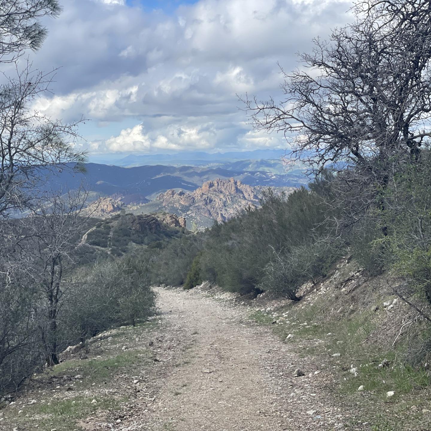 Rocky trail heading down chaparral covered mountain with rock formations seen from a distance.
