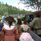 A crowd of people in historic clothing watch a woman lift weighted buckets attached to a yoke.