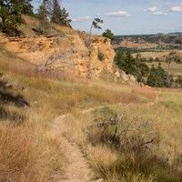 Path winding through prairie grass, with cliff face, red rocks, and pine trees in background.