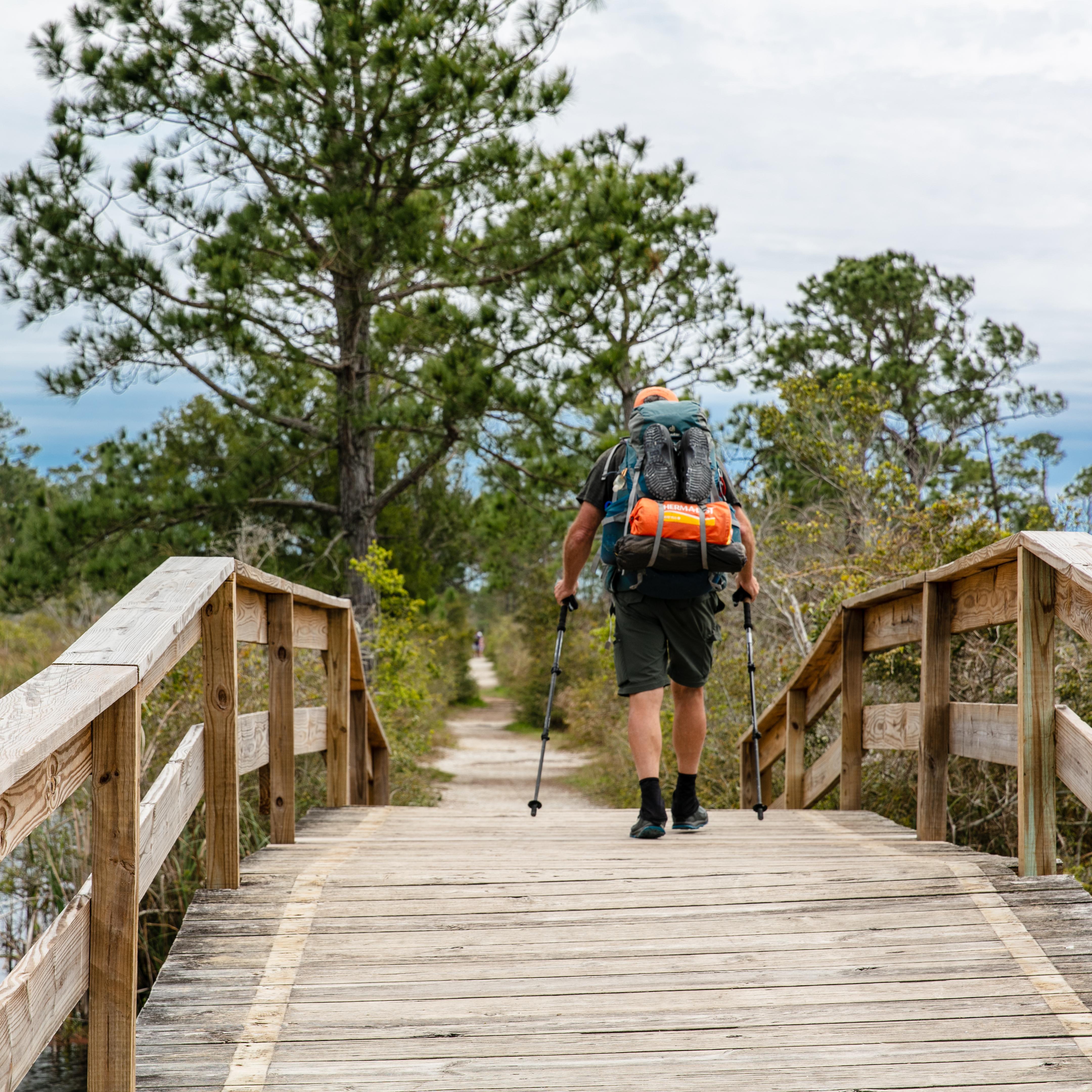 A backpacker crosses over a bridge with poles in hand.