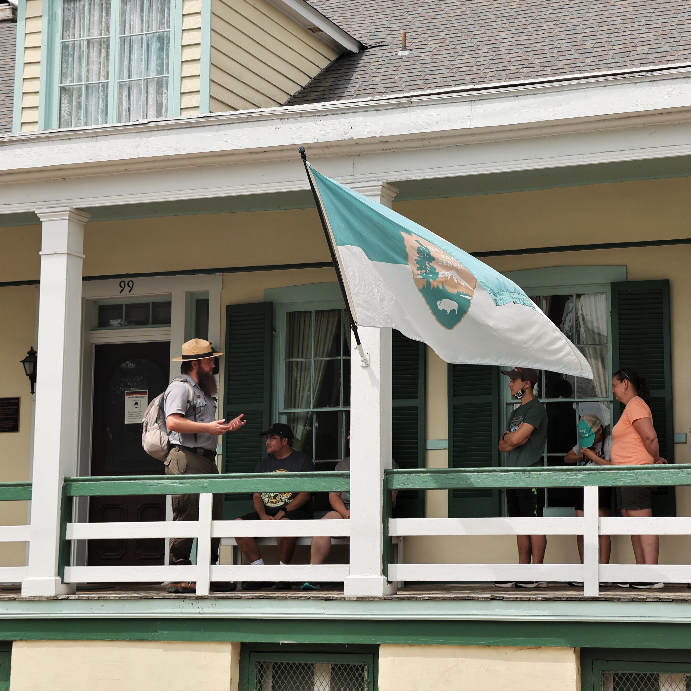 Park ranger talking un a covered porch of a cream house to several people. 