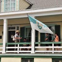 Park ranger talking un a covered porch of a cream house to several people.