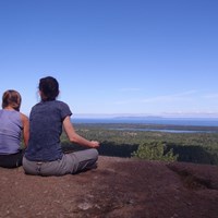 Two people sit on a rocky outcropping over looking a forest and lakes on an island in Lake Superior.