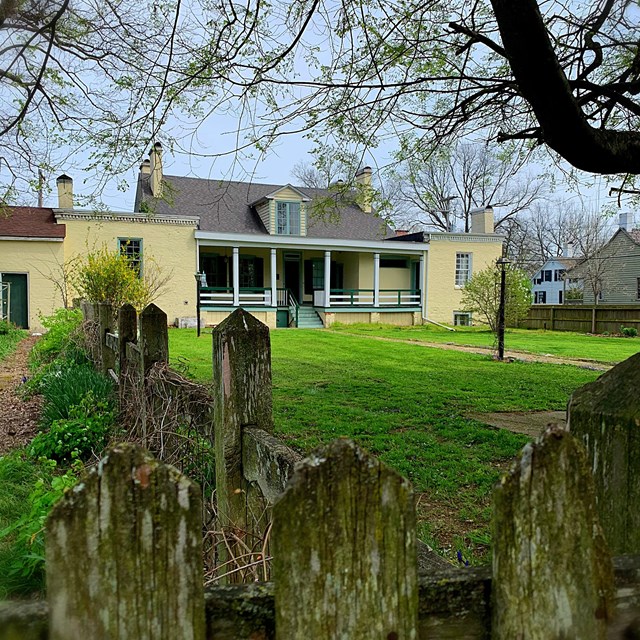 Two story yellow and green house with a covered porch. Grassy lawn and fence posts in the foreground