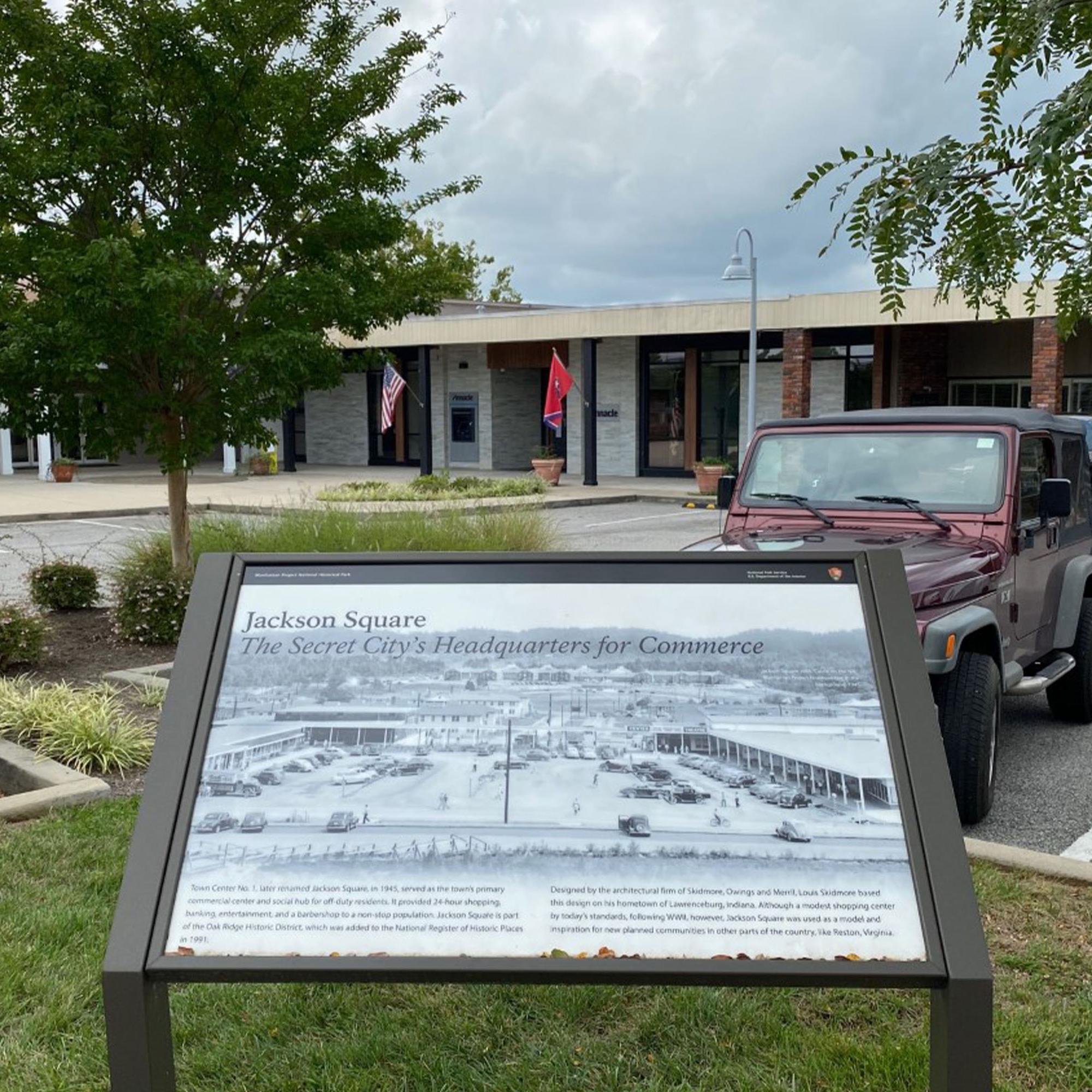 A strip mall with cars parked and historical marker in the foreground.