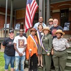 A group of people stand on the steps of the Hotel Florence veranda next to American flag smiling.