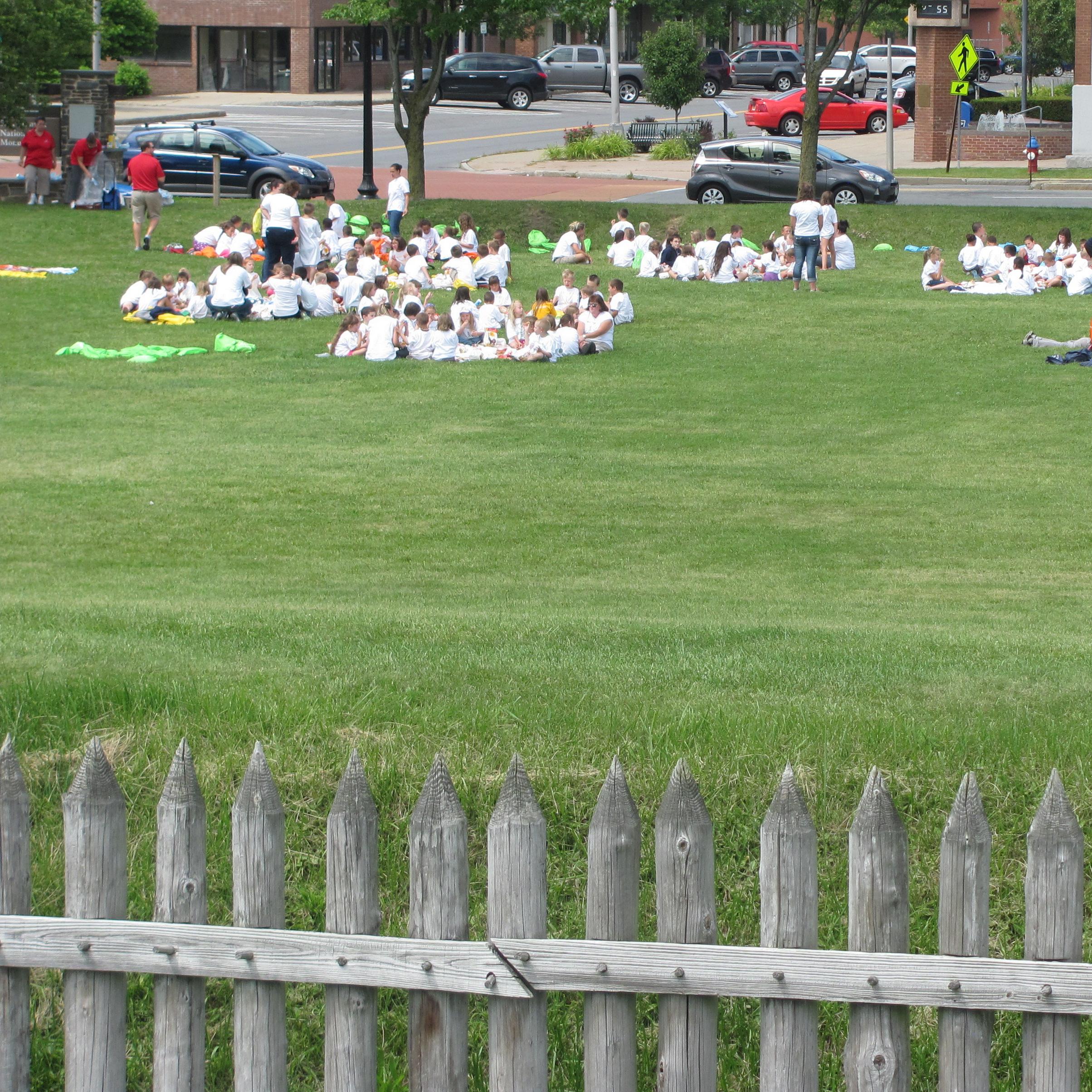 A large group of children sit on blankets scattered across the lawn.