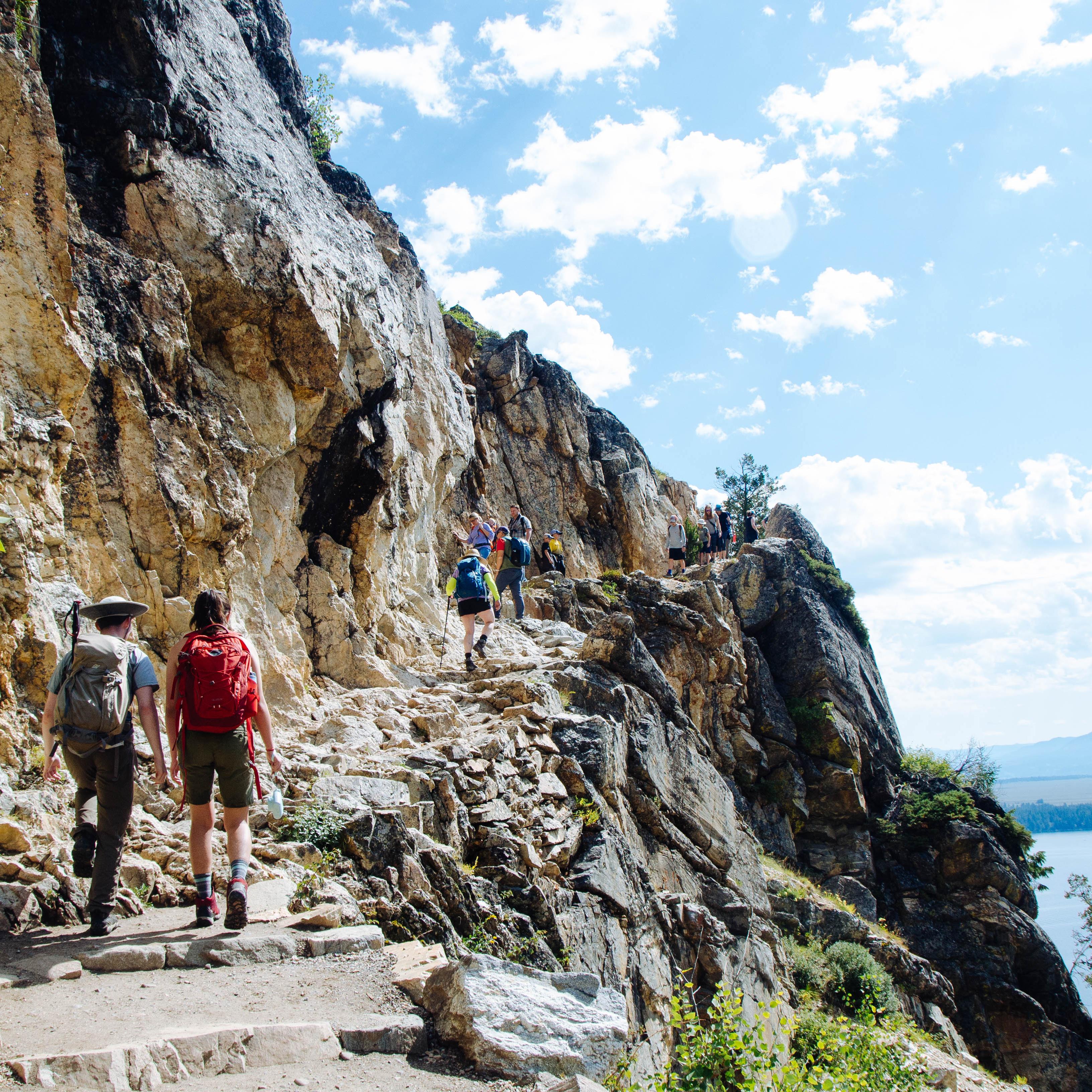 Hikers walk up a rocky trail with a cliff on the right.